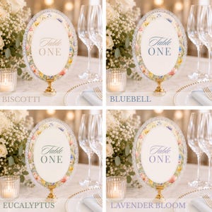 K&ouml;nnte beinhalten: Vier ovale Tischnummerkarten mit der Aufschrift Table ONE, jeweils mit floralem Rand und gewelltem Abschluss, montiert auf einem goldfarbenen Metallst&auml;nder. Die Designs umfassen die Farbpaletten Biscotti, Bluebell, Eucalyptus und Lavender Bloom.