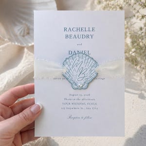 Op de afbeelding: Een trouwkaart met de namen Rachelle Beaudry en Daniel, met een schelpmotief met de initialen "MB". De uitnodiging bevat de datum 15 augustus 2028 en details van de locatie, vastgebonden met een wit lint.