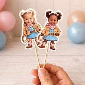 K&ouml;nnte beinhalten: Zwei Cupcake-Topper mit Illustrationen von jungen M&auml;dchen in Cowboy-Outfits. Jedes M&auml;dchen tr&auml;gt ein blaues Kleid, eine beige Jacke, ein rosa Halstuch und Cowboy-Stiefel. Sie halten ein Lasso und haben wei&szlig;e Schleifen im Haar. Die Topper sind an Holzst&auml;bchen befestigt.