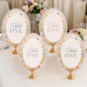 K&ouml;nnte beinhalten: Vier ovale Tischnummernschilder mit der Aufschrift Table ONE, jeweils mit einem Blumenrand aus rosa, gelben und blauen Wildblumen, einer wei&szlig;en Mitte und einem wei&szlig;en Spitzenrand. Jedes Schild steht auf einem kleinen, runden, goldfarbenen Metallst&auml;nder.