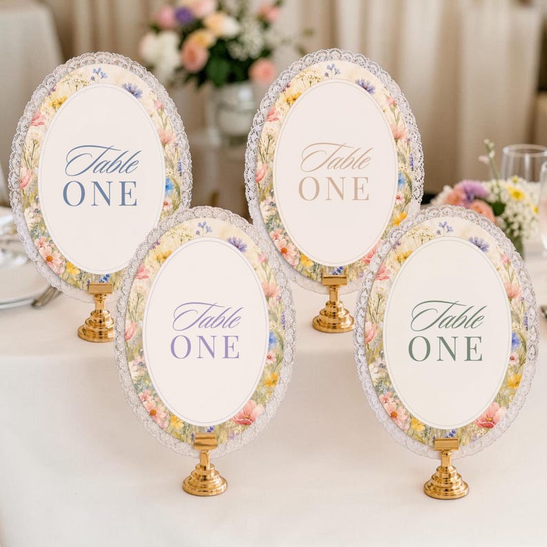 K&ouml;nnte beinhalten: Vier ovale Tischnummernschilder mit der Aufschrift Table ONE, jeweils mit einem Blumenrand aus rosa, gelben und blauen Wildblumen, einer wei&szlig;en Mitte und einem wei&szlig;en Spitzenrand. Jedes Schild steht auf einem kleinen, runden, goldfarbenen Metallst&auml;nder.