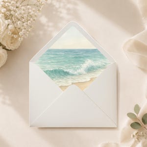 May include: A white envelope displaying a watercolor painting of a beach scene with turquoise water and a breaking wave. The envelope is surrounded by white flowers and a ribbon on a cream-colored surface.