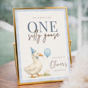 May include: A framed sign with a gold border and a blue plaid background. The sign reads "Celebrating ONE silly goose" with a watercolor illustration of a goose wearing a party hat and holding a balloon. The text "Welcome to Oliver's 1st Birthday" is also displayed.