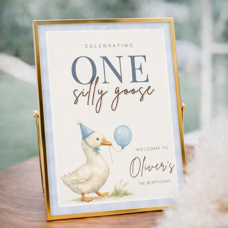 May include: A framed sign with a gold border and a blue plaid background. The sign reads "Celebrating ONE silly goose" with a watercolor illustration of a goose wearing a party hat and holding a balloon. The text "Welcome to Oliver's 1st Birthday" is also displayed.