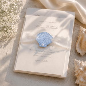 Op de afbeelding: Trouwuitnodigingsset met een stapel cr&egrave;mekleurige kaarten vastgebonden met een transparant lint. Een lichtblauwe schelpvormige versiering met initialen en een parel siert het lint. De tekst luidt "Helena & Brandon" en "Santa Barbara, Californi&euml;."
