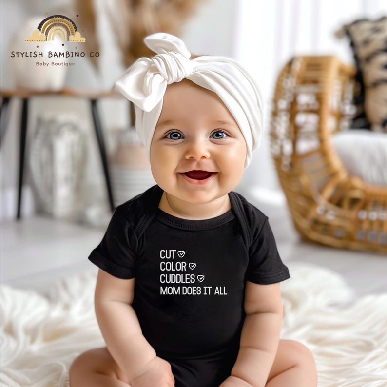 May include: A baby wearing a black bodysuit with the text "CUT &hearts; COLOR &hearts; CUDDLES &hearts; MOM DOES IT ALL" and a white turban headband.