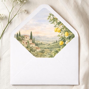Puede incluir: Un sobre blanco con una ilustraci&oacute;n en acuarela de un paisaje toscano visible en el interior. La obra de arte representa un pueblo en la ladera, cipreses y un limonero con frutos amarillos y flores blancas. El sobre est&aacute; sobre una superficie blanca.