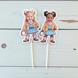 K&ouml;nnte beinhalten: Zwei Cupcake-Topper mit Illustrationen junger M&auml;dchen in Cowboy-Outfits. Jedes M&auml;dchen tr&auml;gt ein blaues Kleid, Cowboy-Stiefel, ein rosa Halstuch und eine Jacke mit Fransen. Ein M&auml;dchen ist blond, das andere hat dunkle Haare.
