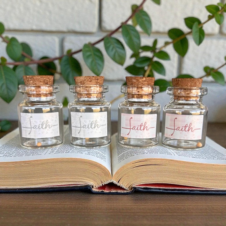 May include: Four small glass jars with cork stoppers, each labelled with the word "Faith" on a white or pink label. The jars are arranged on an open book, with green leaves and a white brick wall in the background.