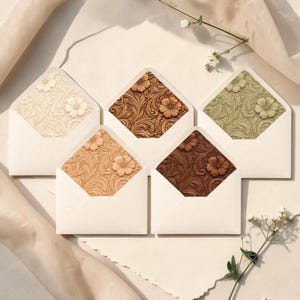 May include: Five white envelopes with floral patterned liners in various colors: cream, brown, tan, dark brown, and olive green. Each envelope has a flower design in the center of the liner. The envelopes are arranged on a light-colored surface.