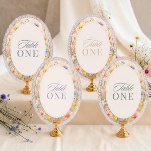 K&ouml;nnte beinhalten: Vier ovale Tischnummernschilder mit der Aufschrift Table ONE, jeweils mit einem Blumenrand aus rosa, gelben und blauen Wildblumen, einer wei&szlig;en Mitte und einem Spitzenrand. Jedes Schild steht auf einem kleinen, runden, goldfarbenen Metallst&auml;nder.