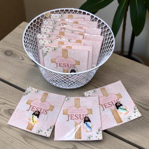 May include: A white wire basket filled with pink square cards featuring a cross, heart, and the words "EVERYONE NEEDS A LITTLE JESUS." Several cards are outside the basket, with a Jesus figurine.