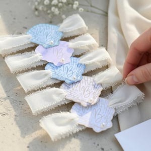 Op de afbeelding: Een verzameling schelpvormige labels in pastelblauw, lavendel en wit, elk versierd met de initialen "MB" en een kleine parel. De labels zijn gecombineerd met delicate, gerafelde witte linten, wat een zachte, elegante uitstraling cre&euml;ert.