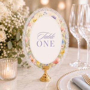 K&ouml;nnte beinhalten: Ein ovales Tischnummernschild mit einem Blumenrand aus rosa, gelben und blauen Wildblumen, umrahmt von wei&szlig;er Spitze. Die Mitte ist wei&szlig; und tr&auml;gt den violetten Schriftzug Table ONE. Das Schild steht auf einem kleinen goldfarbenen Metallst&auml;nder.