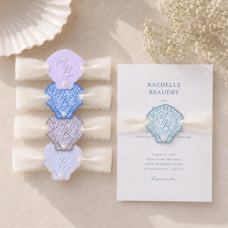 Op de afbeelding: Bruiloftsuitnodigingsset met een schelpmotief. De uitnodigingskaart is wit met lichtblauwe tekst en een schelpgrafiek. De set bevat schelpvormige versieringen in pastelkleuren, elk met de initialen "MB" en een lint.