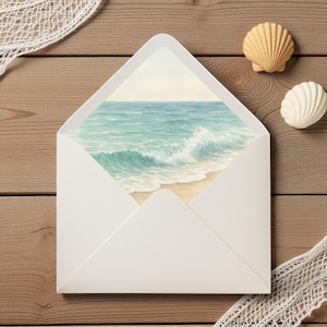 May include: A white envelope with a watercolor painting of a beach scene. The painting shows turquoise waves crashing on a sandy shore. Two seashells and a piece of white netting are on a wooden surface.
