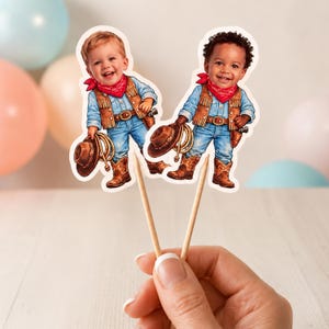 May include: Two cowboy-themed cupcake toppers. Each topper features a smiling toddler dressed in a denim shirt, vest, jeans, cowboy boots, and a red bandana. They hold a cowboy hat and rope. The toppers are attached to wooden sticks.