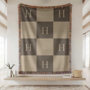 May include: A checkered throw blanket in shades of brown and tan, featuring the letter "H" in each square. The blanket has a fringed edge with a mix of colors. The blanket is hanging on a wall.