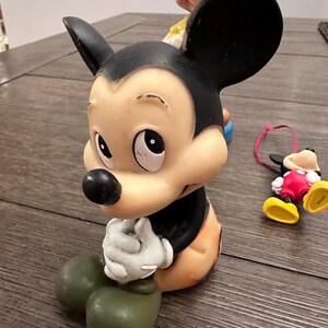 May include: A vintage rubber Mickey Mouse figurine with black ears, a black nose, and a brown body. The figurine is sitting with its hands clasped together and has a sad expression on its face.