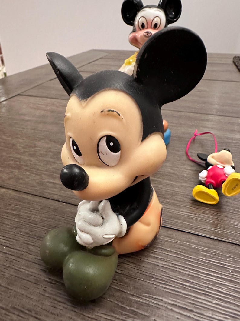 May include: A vintage rubber Mickey Mouse figurine with black ears, a black nose, and a brown body. The figurine is sitting with its hands clasped together and has a sad expression on its face.