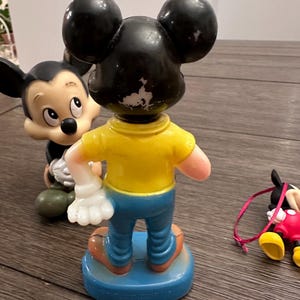 May include: Three vintage plastic Mickey Mouse figurines. One is standing with a yellow shirt and blue trousers. Another is lying down with a red shirt and yellow shoes. The third is standing with a black shirt and white gloves.
