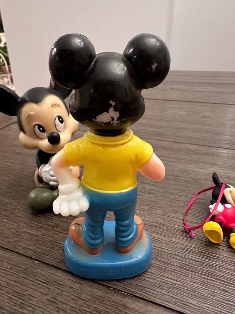 May include: Three vintage plastic Mickey Mouse figurines. One is standing with a yellow shirt and blue trousers. Another is lying down with a red shirt and yellow shoes. The third is standing with a black shirt and white gloves.