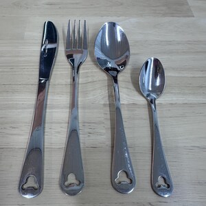 May include: A set of four stainless steel silverware pieces: a knife, fork, and two spoons. Each piece has a teardrop-shaped handle with a decorative cutout at the base. The silverware is arranged on a light brown wooden surface.