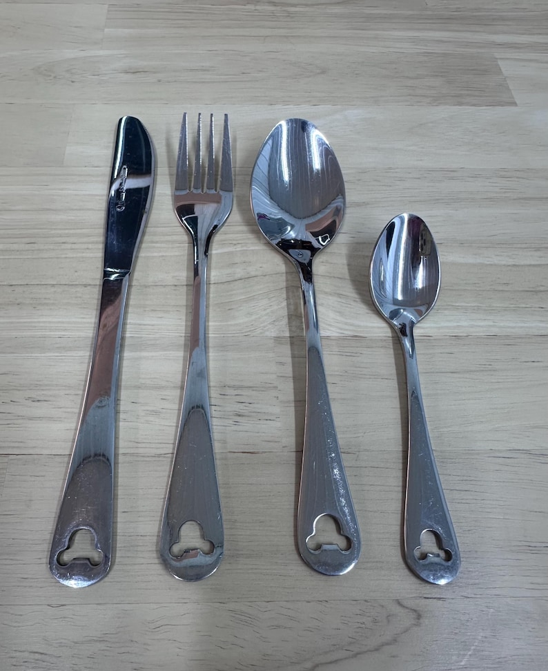 May include: A set of four stainless steel silverware pieces: a knife, fork, and two spoons. Each piece has a teardrop-shaped handle with a decorative cutout at the base. The silverware is arranged on a light brown wooden surface.