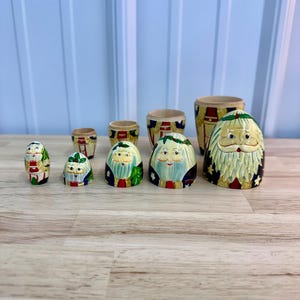 May include: A set of five nesting dolls painted to resemble Santa Claus figures with white beards, red and blue clothing, and holly accents. The set includes five solid wooden doll bases and five matching removable tops, all arranged in a row.