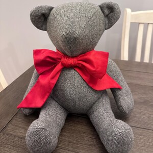 May include: A gray tweed teddy bear with a large red bow. The bear is sitting on a wooden surface.