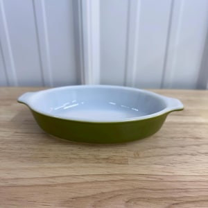 May include: An oval-shaped, olive green and white baking dish with two small handles. The dish has a glossy finish and sits on a wooden surface. The interior is white, and the exterior is a solid olive green.