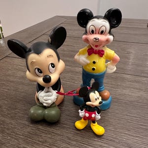 May include: Three vintage Mickey Mouse figurines. The largest figurine is standing with a yellow shirt, blue trousers, and a red bow tie. The middle figurine is sitting with its hands clasped. The smallest figurine is standing with red shorts and black shoes.