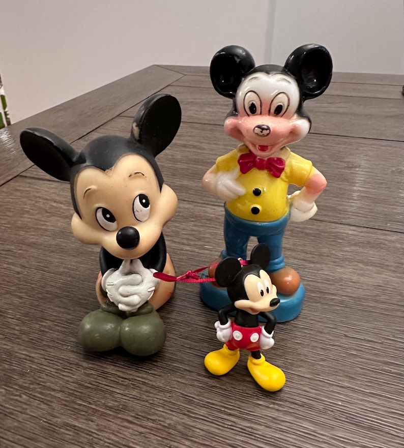 May include: Three vintage Mickey Mouse figurines. The largest figurine is standing with a yellow shirt, blue trousers, and a red bow tie. The middle figurine is sitting with its hands clasped. The smallest figurine is standing with red shorts and black shoes.