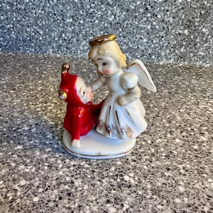 May include: A vintage porcelain figurine depicting an angel in a white dress with gold accents and a halo, interacting with a small figure in a red outfit. The figurine is on a white base and has a classic, detailed design.