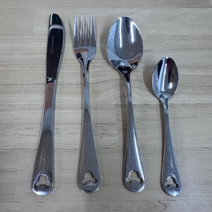 May include: A set of four stainless steel utensils: a knife, fork, spoon, and teaspoon. Each piece has a teardrop-shaped handle with a decorative cutout. The knife has text on the blade.