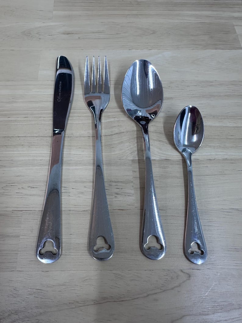 May include: A set of four stainless steel utensils: a knife, fork, spoon, and teaspoon. Each piece has a teardrop-shaped handle with a decorative cutout. The knife has text on the blade.
