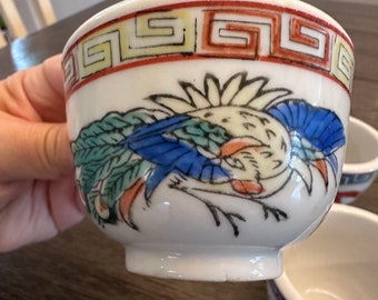 May include: White ceramic bowl with a colorful bird design. The bowl features a red, yellow, and black geometric border. The bird has blue, green, and orange accents. A small, decorative bowl.