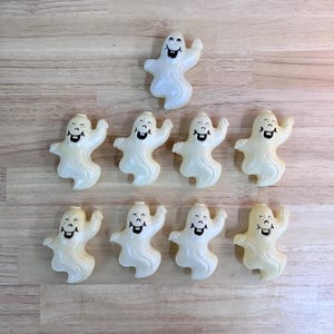 May include: A collection of nine small, off-white plastic ghost figures arranged in two rows. Each ghost features a molded, smiling face with black painted eyes and a wide mouth showing two teeth. The figures have a wavy, flowing base design.