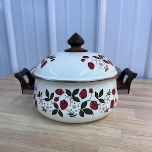 May include: A white enamel pot with a lid, featuring a strawberry and floral design. The pot has dark wooden handles and a matching wooden knob on the lid. The pot is sitting on a wooden surface.
