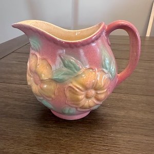 May include: A pink ceramic pitcher with a curved handle and spout. The pitcher features embossed floral designs in shades of yellow, peach, and green. The interior is a light yellow color.