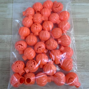 May include: A clear plastic resealable bag filled with numerous small, bright orange plastic pumpkin-shaped light covers. Each pumpkin features a carved jack-o'-lantern face with triangular eyes and a mouth, along with a ribbed texture and a circular opening.