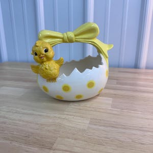 May include: A ceramic Easter basket shaped like a cracked egg, decorated with yellow polka dots. A yellow bow forms the handle, and a yellow chick is perched on the side. The basket is on a wooden surface.