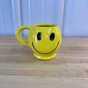 May include: A yellow ceramic mug with a smiley face design. The mug has a rounded shape, a handle, and a black smiling mouth and eyes. The mug is sitting on a wooden surface.