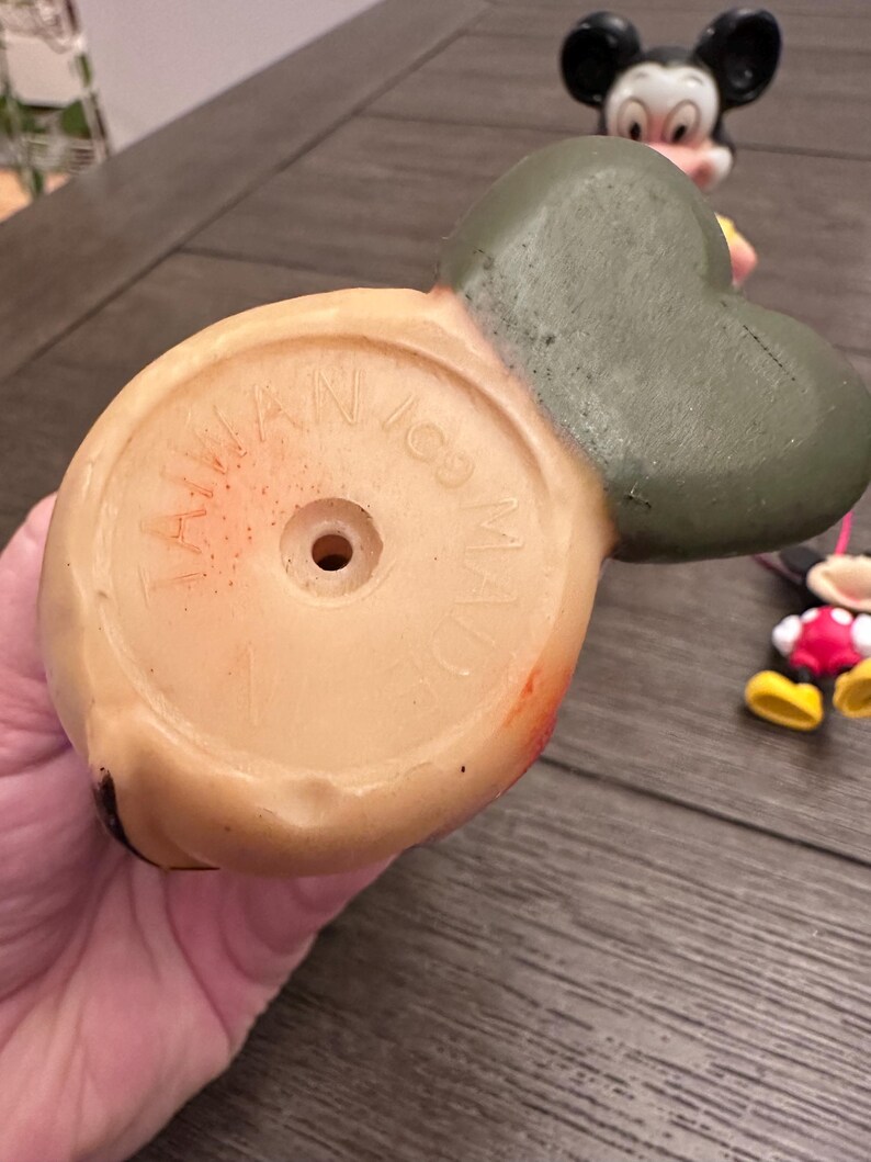 May include: Vintage rubber Mickey Mouse toy with a green hat and red shoes. The bottom of the toy has a circular base with the text "Walt Disney Made in Japan" printed on it.