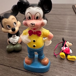May include: Three vintage Mickey Mouse figurines. The largest figurine is standing with a yellow shirt, blue trousers, and a red bow tie. The other two figurines are smaller and are sitting. One is black and white and the other is red and black.