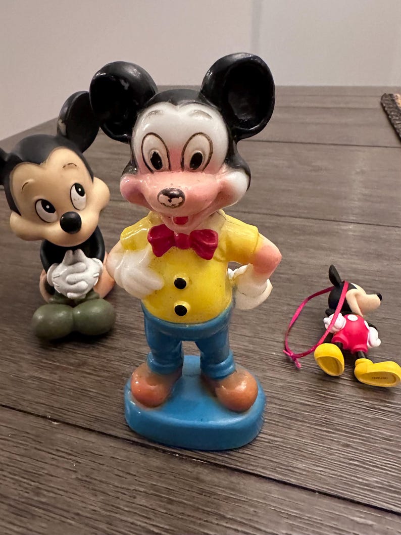 May include: Three vintage Mickey Mouse figurines. The largest figurine is standing with a yellow shirt, blue trousers, and a red bow tie. The other two figurines are smaller and are sitting. One is black and white and the other is red and black.