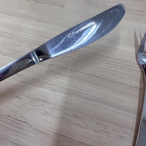 May include: A stainless steel dinner knife with a serrated edge. The knife has a reflective surface and a rounded tip. The handle is smooth and the blade is slightly curved. The knife is part of a silverware set.