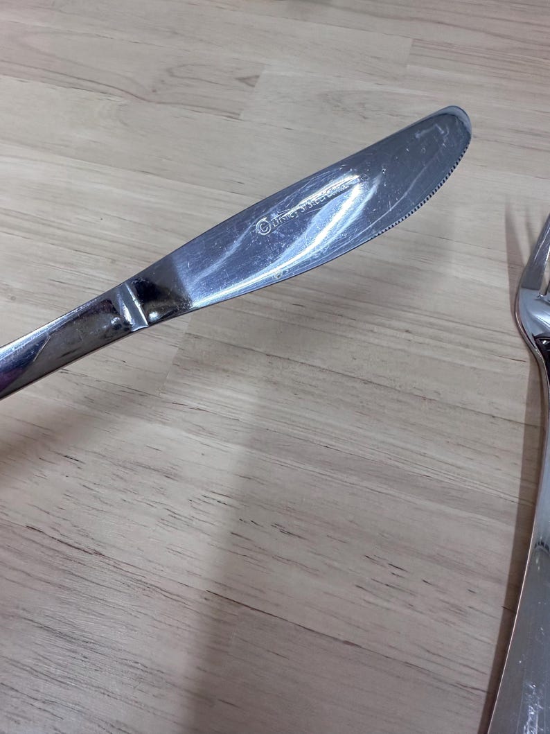 May include: A stainless steel dinner knife with a serrated edge. The knife has a reflective surface and a rounded tip. The handle is smooth and the blade is slightly curved. The knife is part of a silverware set.