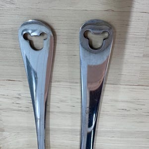May include: Two stainless steel bottle openers. Each opener has a long, slender handle and a head shaped like a cartoon mouse. The openers are reflective and appear to be new.