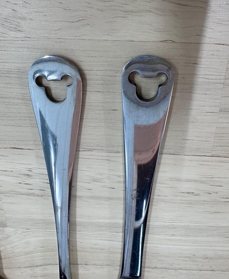 May include: Two stainless steel bottle openers. Each opener has a long, slender handle and a head shaped like a cartoon mouse. The openers are reflective and appear to be new.